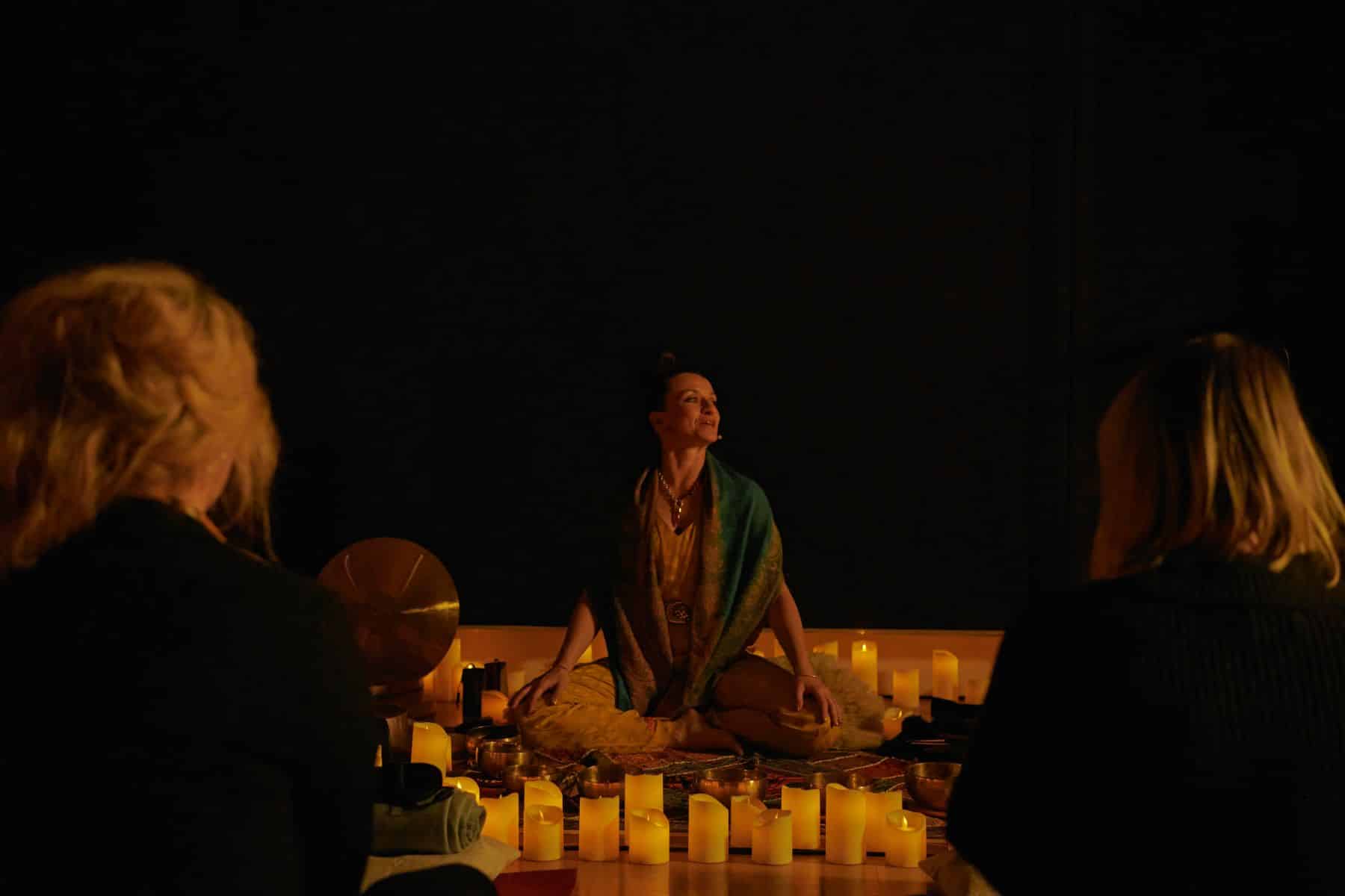 A woman in the center of the image instructing a Mindful Glow class surrounded by candles and two guests watching her.